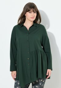 Camicia button-up verde scuro con orlo plissettato, maniche lunghe e colletto; presenta bottoni dorati e una vestibilità leggermente ampia.