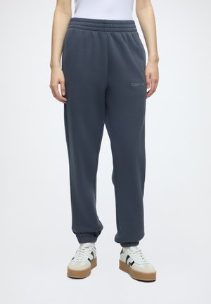 Person iført mørkegrå Calvin Klein joggingbukser med elastiske manchetter og hvide sneakers med sorte detaljer mod en ensfarvet lys baggrund.
