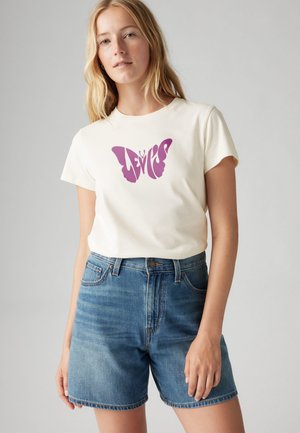 Cremefarbenes T-Shirt mit einem lila Schmetterlingsgrafik, kombiniert mit blauen Jeansshorts. Der Stoff wirkt weich und lässig.