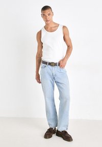 White tank top, light blue denim jeans, gray belt with decorative buckle, and dark brown shoes. Model stands against a plain background.