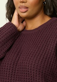 Burgunderfarbener Strickpullover mit einem strukturierten Muster, Rundhalsausschnitt und gerippten Bündchen. Nahaufnahme mit Fokus auf die Stoffdetails und die Form.