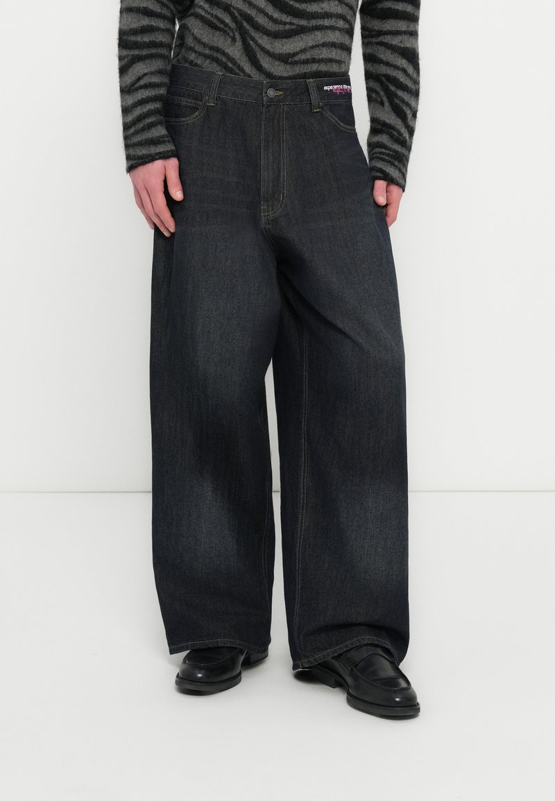 Farmerke tamnog trapera širokih nogavica s visokim strukom, klasičnog dizajna s pet džepova i suptilnim izblijedjenjem. U kombinaciji s crnim cipelama.