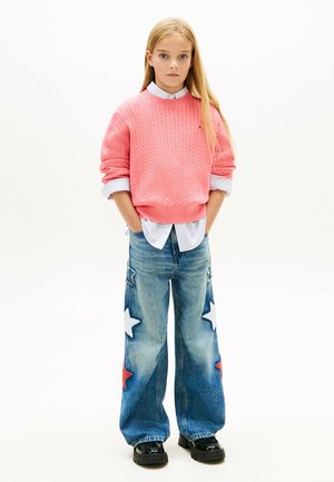 Ragazza in piedi con le mani nelle tasche, indossa un maglione lavorato a maglia rosa, una camicia bianca, jeans larghi con toppe a forma di stella e scarpe nere su sfondo bianco.