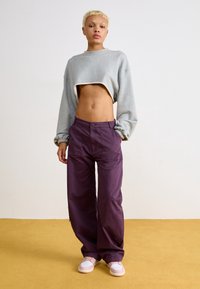 Šedá krátká mikina, volné fialové kalhoty s předními kapsami a světle růžové tenisky na žluté podlaze. Hladká textura, ležérní design.