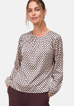Blouse fluide en rose clair avec un motif géométrique couleur bordeaux foncé. Elle a des manches longues et un col froncé. Texture de tissu douce.