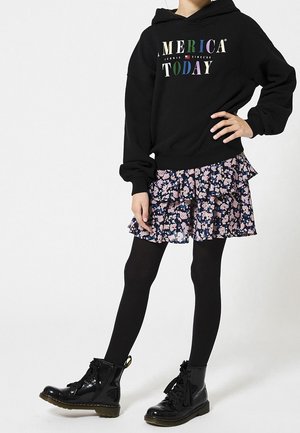 Sudadera negra con "AMERICA TODAY" impreso en letras multicolores, combinada con una falda en capas floreada en azul oscuro y rosa, medias negras y botas negras brillantes.