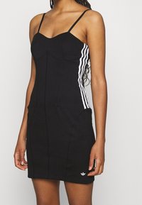 Black fitted dress with thin straps, structured bodice, white three-stripe accents on sides, and small logo at the hem. Stretchy material.