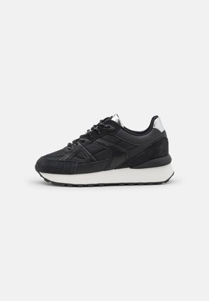 Czarny sneaker z tkaniną o fakturze i panelami z zamszu, biała gruba podeszwa, czarne sznurowadła oraz odblaskowy srebrny detal na pięcie.