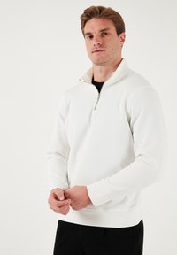 Hanorac alb din fleece cu fermoar, cu mâneci lungi, având un guler înalt și o textură netedă, purtat peste o cămașă neagră.