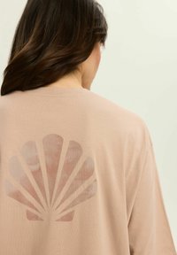 Jasno beżowa koszulka z dużym, wyblakłym wzorem muszli w jasnoróżowym kolorze na plecach. Materiał wydaje się gładki i ma luźny krój.