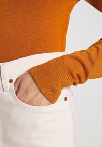 Oranges Strickpullover mit gerippten Bündchen, kombiniert mit weißen Denim-Hosen, die eine Fronttasche und Kupferknopfdetails aufweisen.