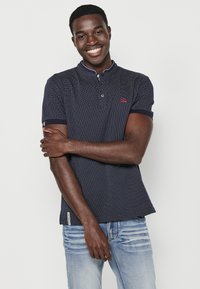 Žydra trumpomis rankovėmis polo marškinėliai su tekstūruotu raštu, kontrastinga apykakle ir mažo logotipo detale. Suderinta su šviesiai mėlynomis džinsais.