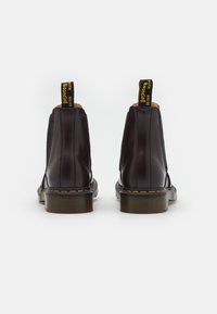 Par de botas de cuero marrón oscuro con costuras amarillas y pestañas negras en el talón que dicen "WITH SOLES Bouncing", vistas desde la parte posterior sobre fondo blanco.