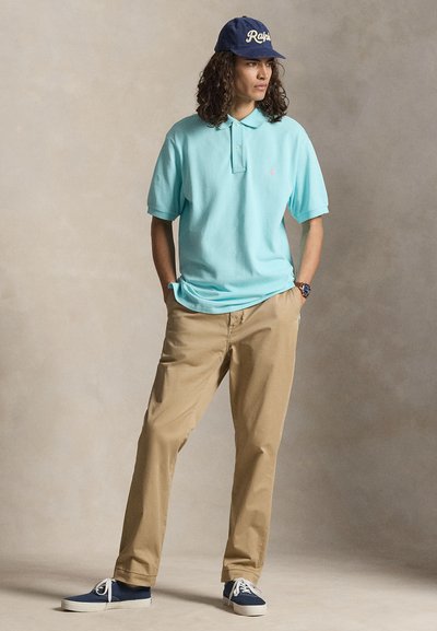 Junger Mann mit langen lockigen Haaren, der eine blaue Kappe, ein hellblaues Poloshirt, beige Hosen und blaue Sneakers trägt und mit den Händen in den Taschen steht.