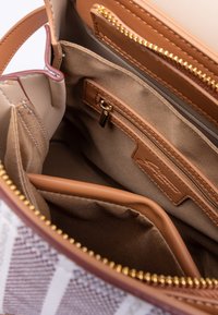 Interno di una borsa beige con scomparti; presenta una tasca con zip e una morbida fodera in tessuto. Include un inserto rimovibile marrone e accessori in oro.