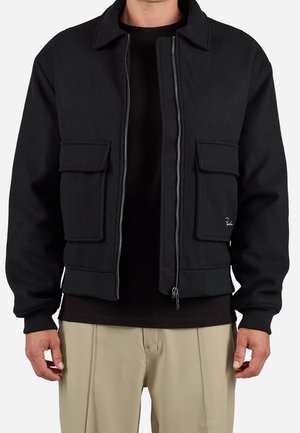 Mann trägt schwarze Jacke mit Reißverschluss und zwei großen vorderen Klappentaschen, schwarzes Hemd und beige Hose, gezeigt vom Hals bis zur Hüfte.