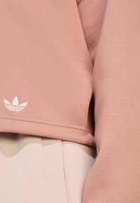 Pogled od blizu na prašnjavi roza rub dukserice s bijelim Adidas Trefoil logotipom iznad bež nabranih hlača.
