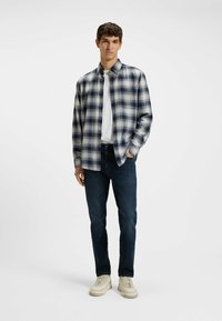 Camisa de franela a cuadros en azul marino y gris, con cuello, abotonada, combinada con jeans azul oscuro y zapatillas crema, de pie sobre un fondo neutro.