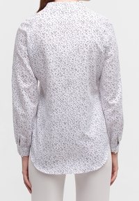 Chemise blanche à motif de petits points noirs dispersés, design col avec manches longues. Comprend un ourlet arrondi et des poignets boutonnés. Texture de tissu lisse.