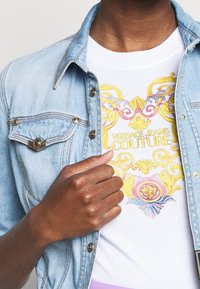 Dżinsowa kurtka z metalowymi detalami w kolorze srebrnym noszona na białej koszulce z kolorowym, ozdobnym wzorem graficznym oraz napisem "VERSACE JEANS COUTURE".