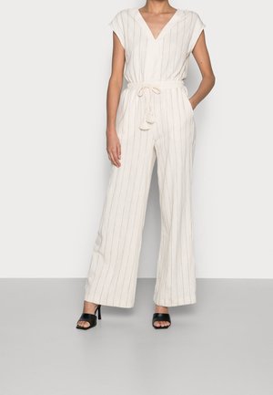 Vrouw draagt een mouwloos ecru jumpsuit met verticale strepen en een taille met trekkoord, gecombineerd met zwarte openhakken.