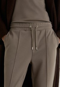 Világosbarna, laza szabású nadrág, húzózsinóros derékrésszel, ráncolásokkal és oldalzsebekkel. Sima anyag enyhe fényű felülettel, hozzáillő felsővel párosítva.