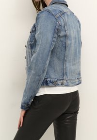 Ljusblå denimjacka med klassisk skärning, med knäppt front, två sidofickor och urblekt struktur. Buren över en vit skjorta.