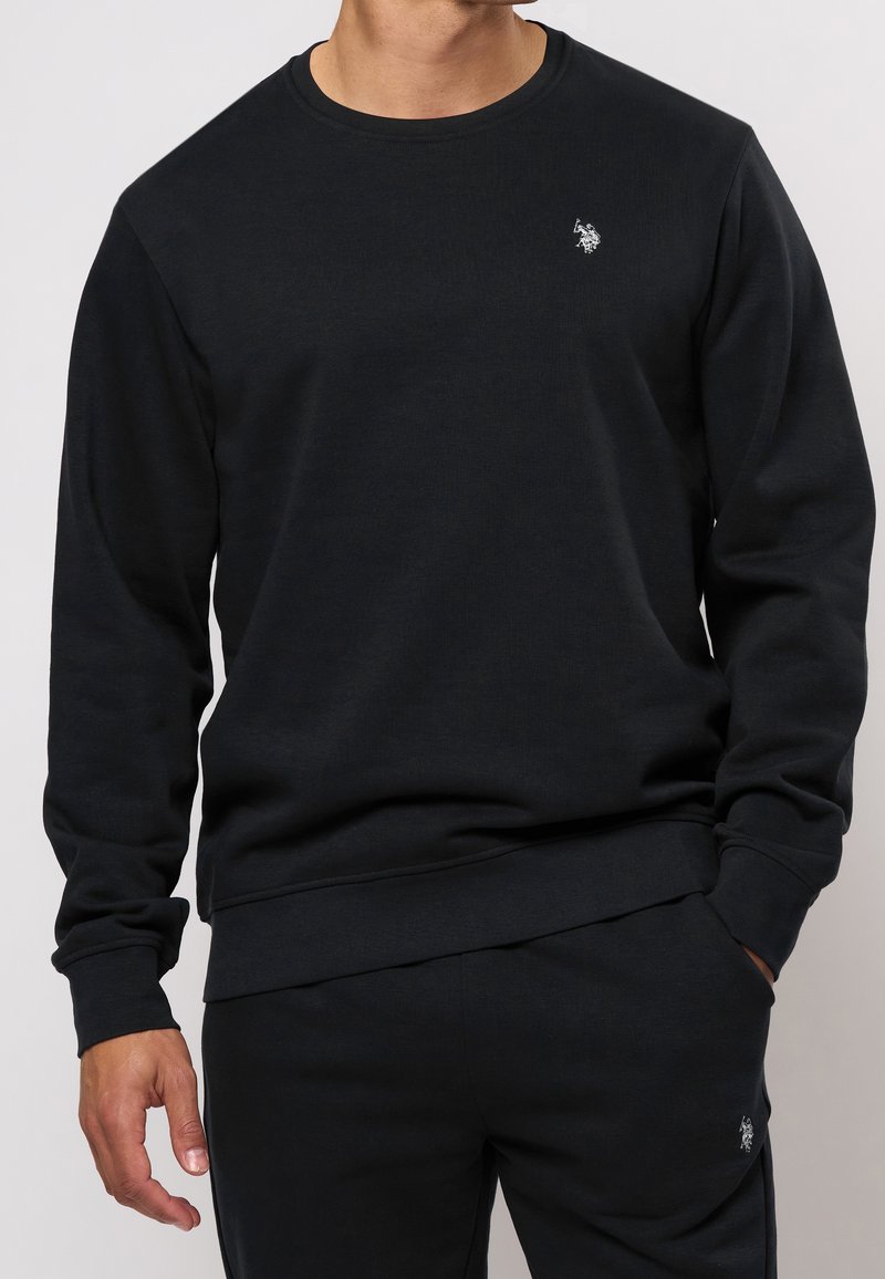 Homme portant un sweat-shirt noir ajusté et un pantalon noir assorti, chacun avec un petit logo brodé blanc d'un joueur de polo sur la poitrine et la cuisse.