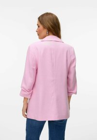Blazer rosa con mangas tres cuartos, corte entallado, textura sutil y cuello con muesca. Llevado sobre vaqueros azules, fondo liso.