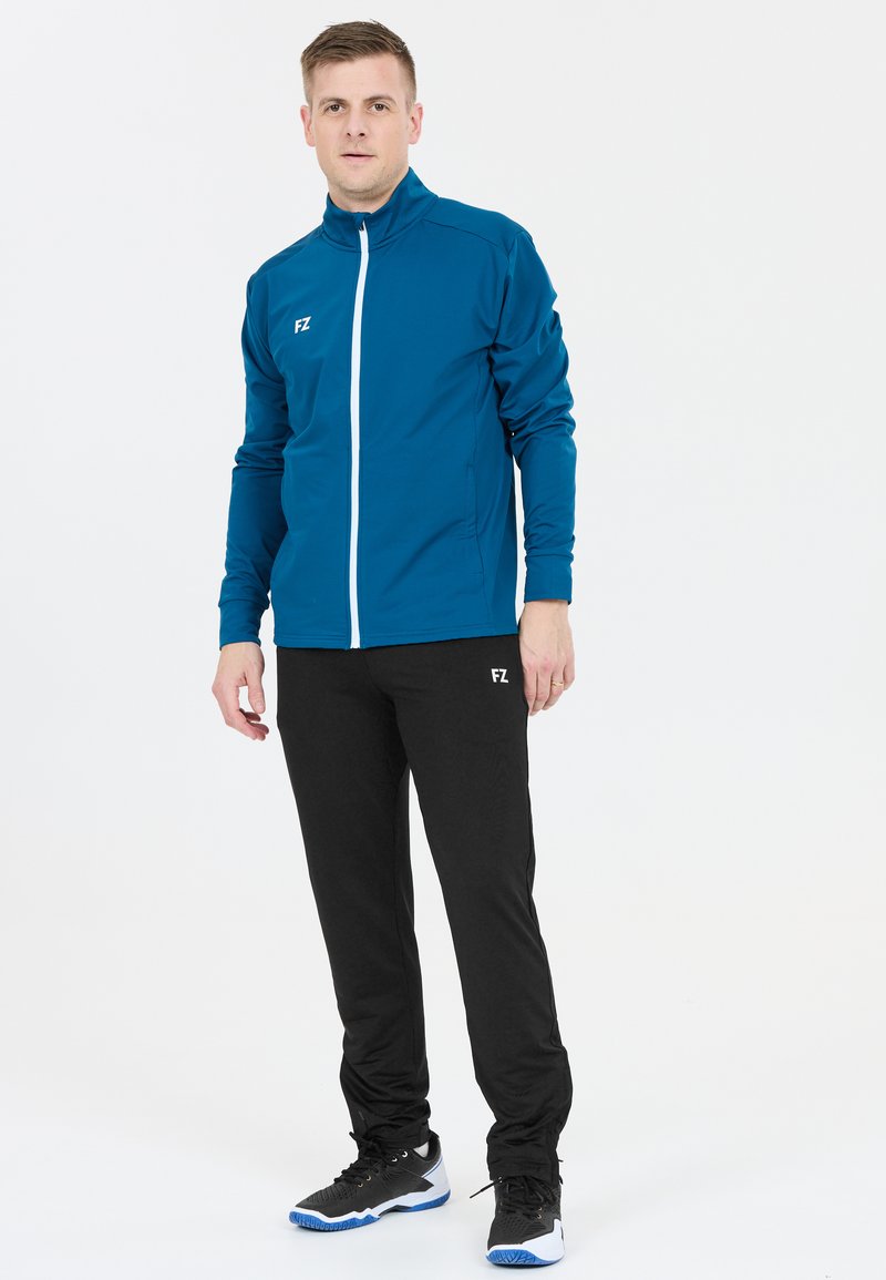 Blå idrottsjacka med hög krage och vit dragkedja, kombinerad med svarta byxor med en logotyp. Modell som bär svarta och blå sneakers.