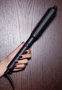 Hand die een zwarte elektrische hete lucht haarborstel vasthoudt, met borstelharen en een aan/uit-knop, tegen een donkere houten achtergrond.