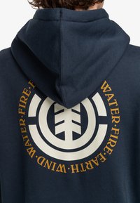 Námornícky modrá mikina s fleece textúrou. Obsahuje veľké kruhové logo v krémovej a zlatej farbe s nápisom "VODA, OHEŇ, ZEM, VZDUCH" okolo neho.