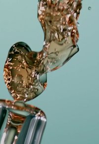 Un liquide transparent aux teintes dorées coule en une forme sculpturale, réfléchissant la lumière sur un fond bleu doux avec des bulles visibles.