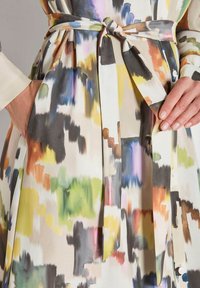 Nahaufnahme von Händen, die einen Gürtel an einem mehrfarbigen Kleid mit abstrakten Mustern und pinselstrichartigen Designs in Gelb, Grün, Schwarz und Blau anpassen.