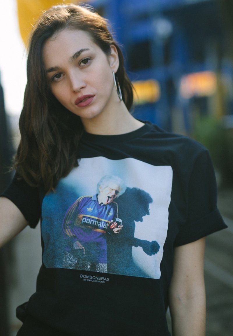 T-shirt nera in cotone con una grafica colorata di un uomo anziano in tuta blu che tiene una bevanda, con la scritta "BOMBONERAS" stampata sotto.