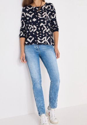 Kvinde iført en marineblå mønstret langærmet top, lyseblå skinny jeans og hvide sneakers med røde detaljer, stående op ad en hvid væg.