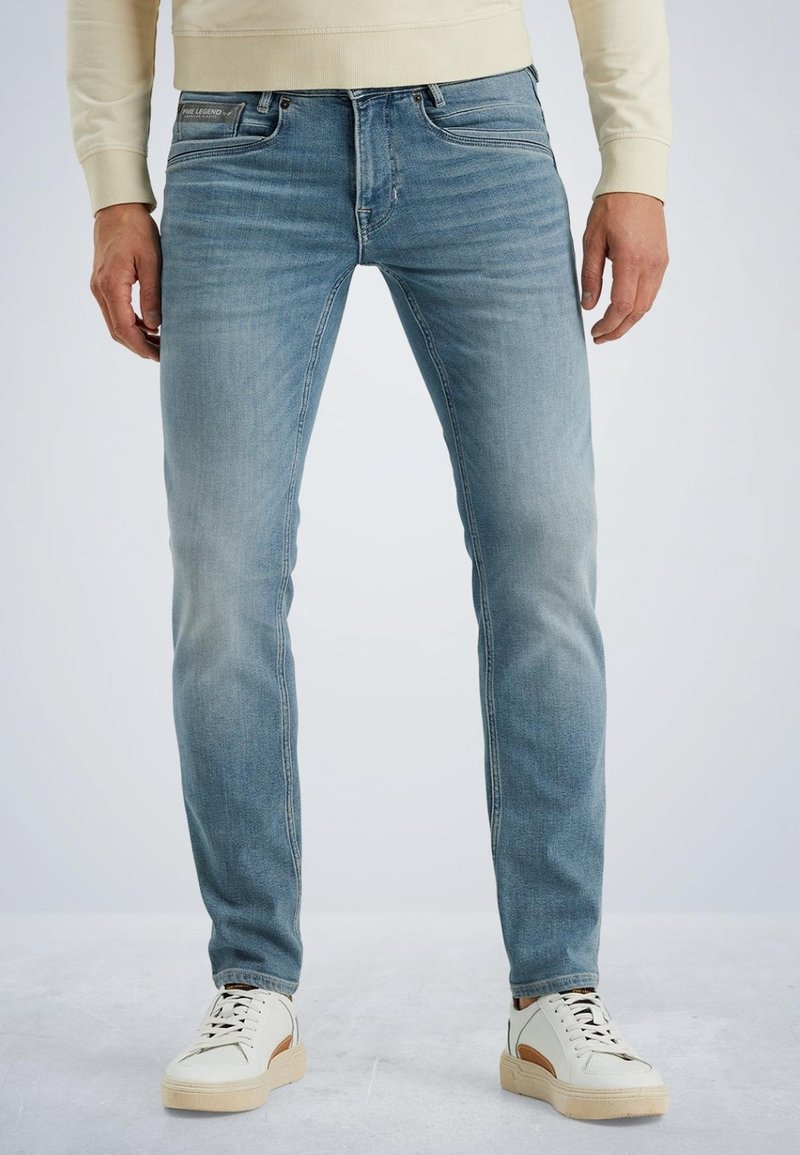Light blue denim jeans with a slim fit, subtle fading, and standard pockets. Worn with white sneakers featuring tan accents.