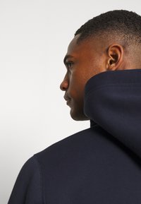 Sudadera con capucha de color marino, con una textura suave, corte relajado y detalles de costura visibles, presentada de lado con un enfoque en la capucha.