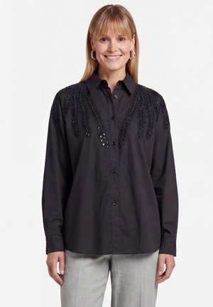 Lachende vrouw met stijl blond haar draagt een zwart overhemd met knopen en gestructureerde bladmotieven op de schouders en lichtgrijze broek.