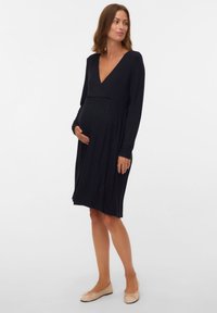 Czarna, długorękawowa sukienka ciążowa z głębokim dekoltem w serek i marszczonym stanem, wykonana z prążkowanej tkaniny. Stylizację dopełniają płaskie beżowe baleriny.