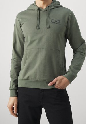 Mężczyzna w oliwkowej bluzie z kapturem z logo EA7 Emporio Armani na piersi, z przednią kieszenią, oraz w czarnych spodniach, ręka w kieszeni.