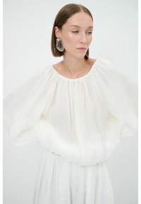 Weiße, leichte Bluse mit rundem Ausschnitt und voluminösen Ärmeln, die eine weiche Textur und geraffte Details aufweist.