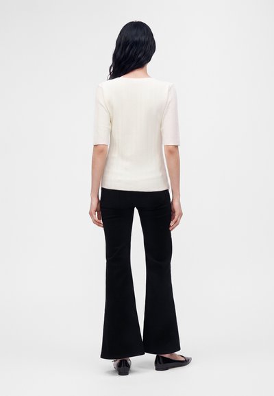 Femme aux longs cheveux foncés, debout de dos, portant un haut blanc à manches courtes, un pantalon noir évasé et des chaussures plates noires.