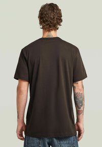 T-shirt in cotone marrone scuro, con maniche corte, taglio diritto e collo rotondo. Presenta una texture liscia e un design semplice sul retro.