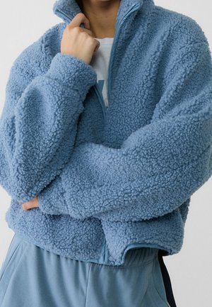 Osoba ubrana w jasnoniebieską, puszystą kurtkę zapinaną na zamek z teksturowanej tkaniny oraz dopasowane spodnie, stojąca z założonymi ramionami na tle o neutralnym kolorze.