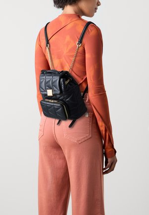Gepolsterter schwarzer Leder-Rucksack mit einer Reißverschluss-Vordertasche, Ketten- und Gurtakzenten; getragen mit einem orangefarbenen Langarmoberteil und rosa Hosen.