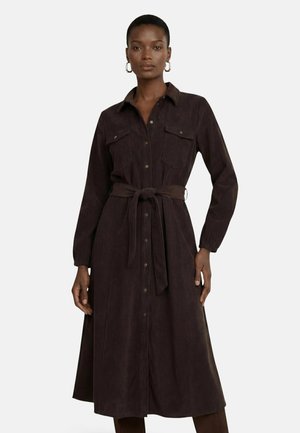 Woman wearing a long-sleeve dark brown button-up dress with chest pockets and a tied belt at the waist, standing against a plain background.