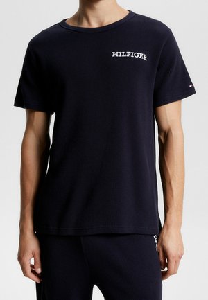 Mand iført en sort tekstureret kortærmet skjorte med "HILFIGER" trykt i hvidt på brystet og et lille logo på venstre ærme.