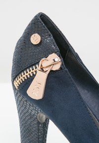 Escarpin bleu marine avec dessus texturé, fermeture éclair métallique avec charme et matière en daim lisse. Motif géométrique près de l'orteil.