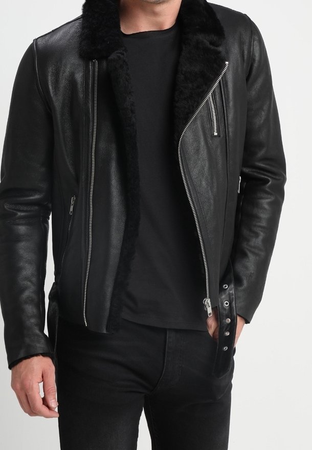 Homme portant une veste en cuir noire avec une doublure en fourrure noire, ouverte sur une chemise noire et un pantalon noir, debout devant un fond uni.